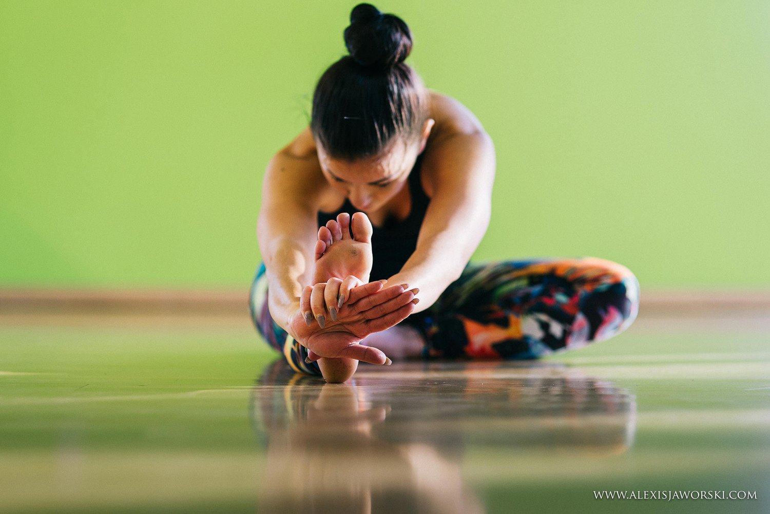 Christie folded forward over one extended leg, hands cupped around the foot, reflected in the polished studio floor.