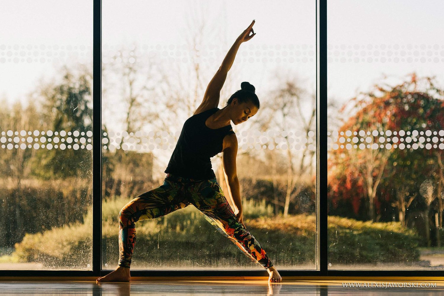 Christie in a wide-legged side bend, one arm lifted, silhouetted against the studio's full-height windows in low golden light.